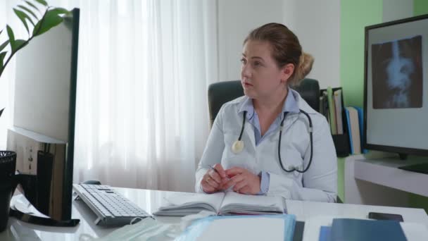 在网上咨询的过程中，女医生通过视频交流与病人进行交流，并在医疗书籍中记录诊断结果，同时坐在X光的办公室背景下 — 图库视频影像