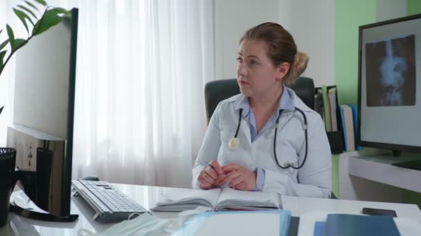 Consulta on-line, médico fêmea se comunica com paciente doente através de comunicação de vídeo e escreve diagnóstico no livro médico enquanto sentado no fundo do escritório de um raio-X — Vídeo de Stock