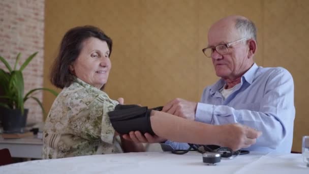 Заботливый старик с помощью монитора артериального давления измеряет давление на здоровую пожилую женщину, сидящую за столом на кухне — стоковое видео