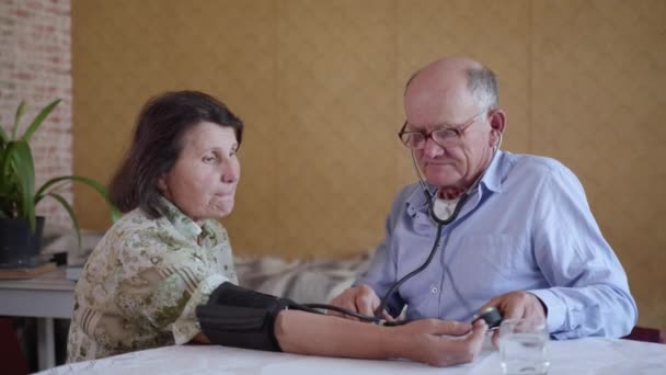 Anciano hombre que cuida comprueba la presión arterial de su vieja esposa con hipertensión usando un monitor de presión arterial mientras está sentado en casa en la mesa — Vídeo de stock