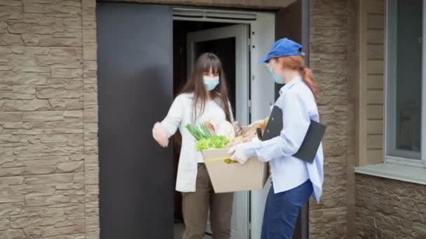 Voedsellevering in quarantaine, jong meisje tijdens isolatie als gevolg van pandemie ontvangt haar bestelling van voedsel door een vrouwelijke koerier met medisch masker, bril en handschoenen ter bescherming tegen virus — Stockvideo