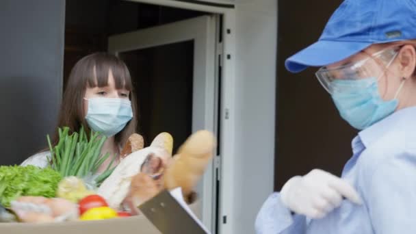 Zakupy online, pracownica kurierska nosząca ochronne rękawice medyczne, okulary i niebieski mundur przynosi żywność zamówioną w sklepie internetowym do domu klientki podczas samodzielnej izolacji oraz — Wideo stockowe