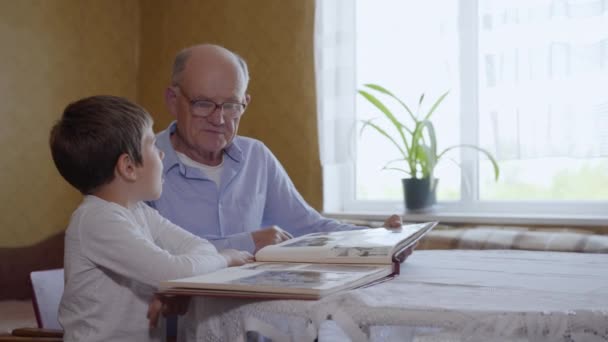 Cariñoso abuelo muestra a su querido nieto un álbum de fotos de la familia sentado en una mesa — Vídeos de Stock