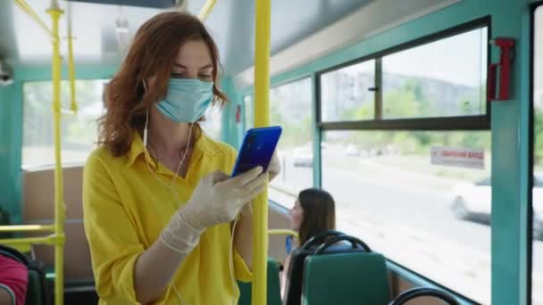 Mladá dívka nosí lékařskou masku a rukavice poslouchá hudbu se sluchátky na svém smartphonu v autobuse, jak cestuje po městě po karanténě byla — Stock video