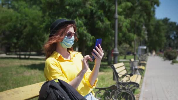 Υγειονομική περίθαλψη, νεαρό χαρούμενο κορίτσι σε ιατρική μάσκα για να προστατεύσει τον εαυτό της από τον ιό και τη μόλυνση και χρησιμοποιεί το κινητό της τηλέφωνο, ενώ χαλαρώνει στο πάρκο στον πάγκο, στη συνέχεια βγάζει αναπνευστήρα και απολαμβάνει φρέσκο αέρα — Αρχείο Βίντεο