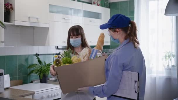 Jovem mensageiro menina em máscara médica e luvas entrega produtos para jovem cliente feminino no respirador para se proteger do vírus e infecção durante a pandemia — Vídeo de Stock