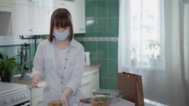Pandemia, mujer joven en máscara médica trata los alimentos entregados a casa con un antiséptico para proteger contra el coronavirus durante la cuarentena, precauciones — Vídeo de stock
