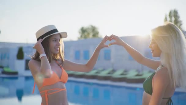 Lifestyle, jonge meisjes in zwemkleding tonen hart teken met de handen op de achtergrond van zonnestralen in de zomer resort — Stockvideo