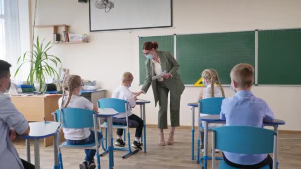 École après quarantaine, professeur masqué distribuant des dépliants avec affectation aux élèves assis aux bureaux pendant une leçon en classe — Video