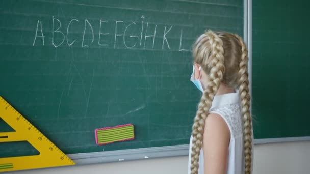 portait of schoolgirl in medical mask writes in chalk the english alphabet in classroom, pupil look on camera near blackboard at school
