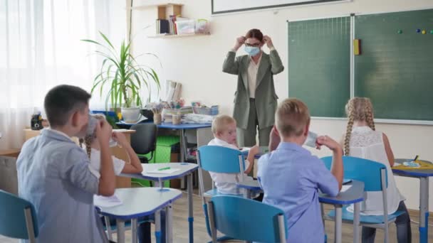 Escolas reabertas, o professor feminino em copos perto do quadro-negro mostra a uns alunos como pôr a máscara médica no rosto antes da lição na sala de aula — Vídeo de Stock