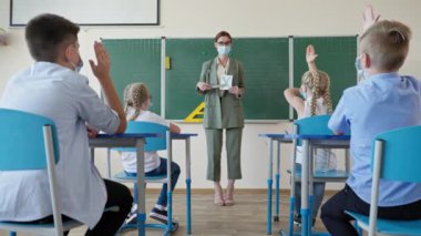 Öğrenci kartlarını sınıfın önünde gösteren maskeli öğretmen. Çocuklar dikkatle dinler ve ders sırasında el kaldırır.