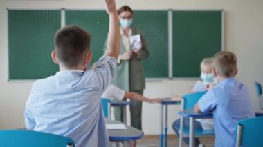 Maskeli okul çocuğu öğretmen sorusuna cevap vermek için elini kaldırıyor. İlkokuldaki bir ders sırasında öğrenciler masalarında oturuyor.