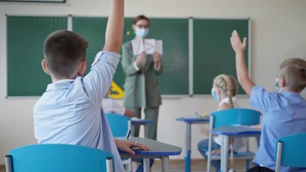 Портрет школьника в маске поднимает руку и отвечает на вопрос учителя, затем поворачивается к камере, показывая большой палец на столе во время урока. — стоковое видео