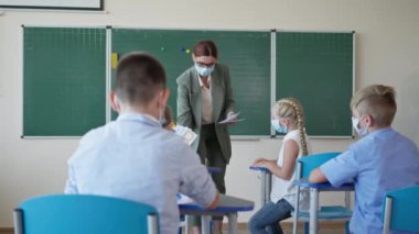 Yeni normallik, ders sırasında masada oturan kartlarla çocuklar, okulda kameraya bakan maskeli mutlu bir öğrenci portresi.