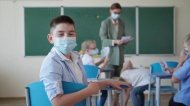 Tıp maskeli öğrencinin portresi. Ders sırasında kameraya bakıyor. Sınıf arkadaşları ve öğretmenlerin arka planında oturuyor.