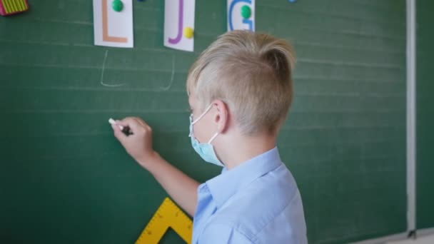 Colegial en máscara médica en la pizarra escribe cartas con tiza en el aula en la escuela después de la cuarentena y el bloqueo — Vídeo de stock