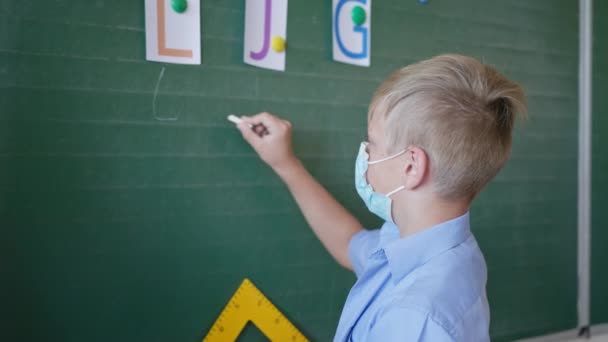 Schooljongen met medisch masker in de buurt van schoolbord schrijft in krijt de brieven in de klas, school na quarantaine en lockdown — Stockvideo