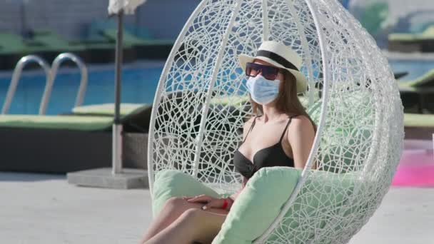 Coronavirus e viaggi, donna in cappello e maschera medica in sedia appesa vicino alla piscina durante le vacanze estive — Video Stock