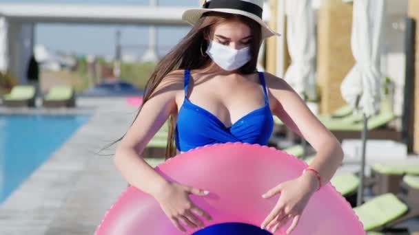 Meisje in stro hoed te houden voorzorgsmaatregelen draagt medisch masker voor bescherming virus en infectie houdt zwemmen ring in zijn handen op de achtergrond van blauw zwembad en ligstoel tijdens de vakantie op zee — Stockvideo