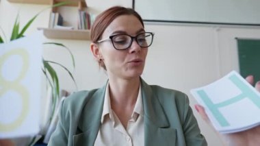 Öğretmen internette kameraya konuşuyor. Gözlüklü genç bir kadının portresi. Elinde kartlar.
