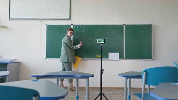 Online onderwijs, pedagoog in bril met liniaal in de buurt van schoolbord met behulp van mobiele telefoon camera opnemen van zichzelf op video tutorial op school — Stockvideo