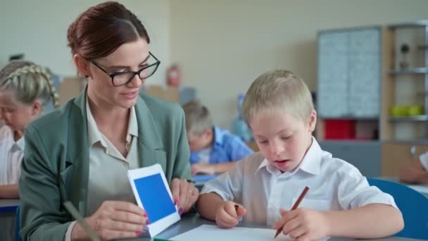 Retrato de niño con discapacidad interactúa con el maestro, alumno aprende formas mientras está sentado con la mujer joven en el escritorio en un aula de la escuela — Vídeos de Stock