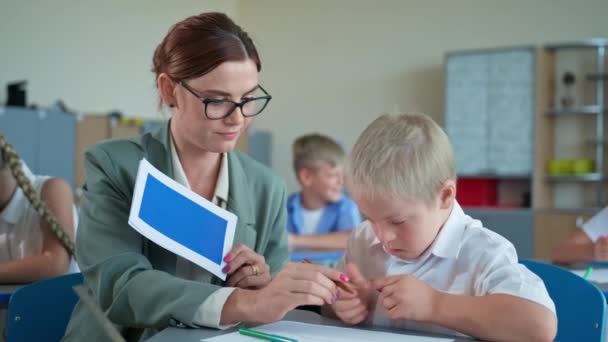 Pojke med Down syndrom i skolan, söt elev med funktionshinder lär sig former medan du sitter med lärare vid skrivbordet i ett klassrum — Stockvideo