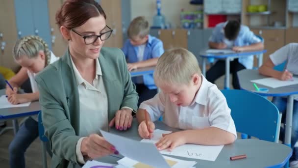 Funktionshindrade pojke med ner syndrom studerar former med lärare sitter vid skrivbordet på bakgrund av klasskamrater under lektionen i skolan — Stockvideo