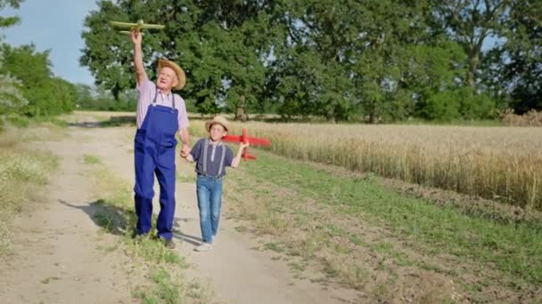 Anciano abuelo junto con su alegre niño varón en sombreros de paja divertirse jugando aviones de juguete — Vídeos de Stock