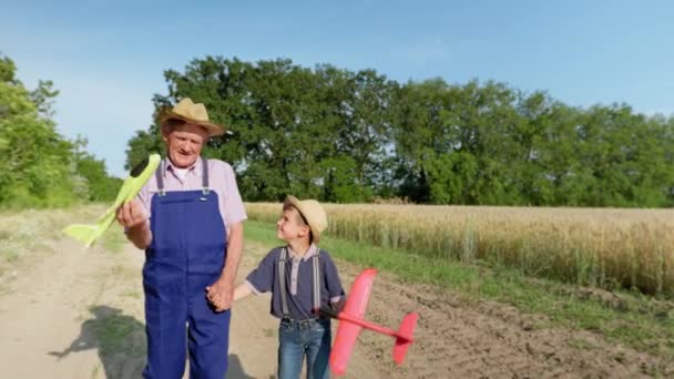 Ευτυχισμένη παιδική ηλικία, αρσενικό παιδί με τον παππού σε ψάθινα καπέλα παίζουν με τα αεροπλάνα παιχνίδι περπάτημα κοντά στο πεδίο — Αρχείο Βίντεο