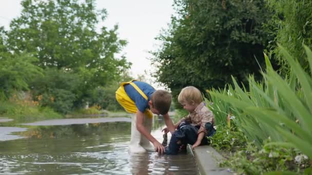 Péče o bratra, roztomilé milující dítě pečující o svého malého a sundá gumové boty nalít vodu při hraní v louži — Stock video