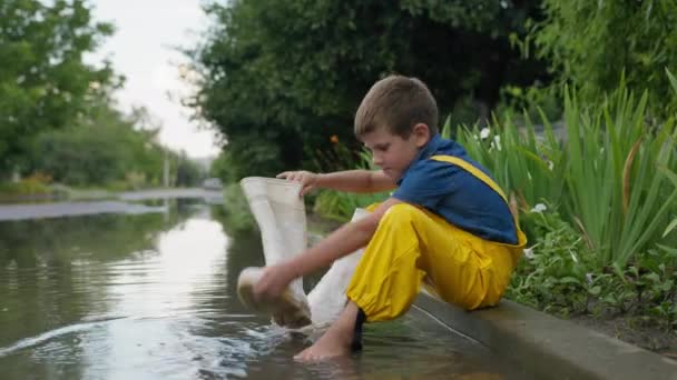 Letnie zabawy, małe szczęśliwe dziecko bawi się w kałuży siedzi na krawężniku i wylewa wodę z gumowych butów na drodze — Wideo stockowe
