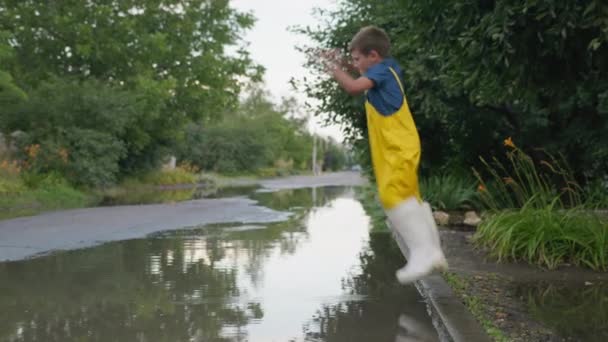 Vacaciones de los niños, niño alegre en botas de goma y monos se divierten saltando de acera en charco por carretera después de la lluvia en el día — Vídeos de Stock