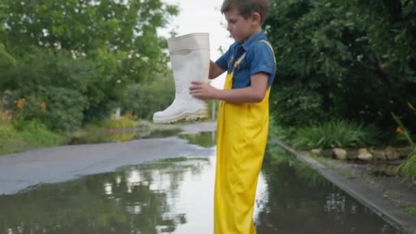 Feliz fin de semana, lindo niño masculino en overoles derrama bota de goma después de divertirse jugando en charco por carretera después de la lluvia con árboles — Vídeos de Stock