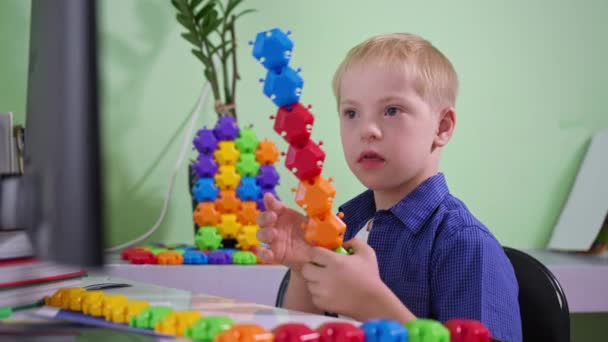 Entretenimento em casa, criança com síndrome de Down está jogando com construtor colorido enquanto sentado à mesa e mostra brinquedos na tela do computador — Vídeo de Stock