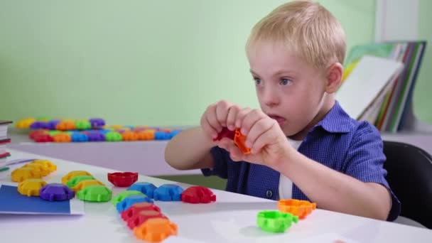 Garoto curioso com síndrome de Down desmonta e brinca com um brinquedo construtor colorido sentado à mesa em casa close-up — Vídeo de Stock