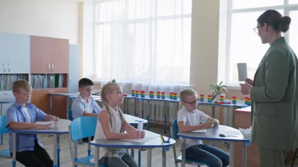 Maestro muestra tarjetas con asignación para los alumnos, los niños levantan las manos sabiendo la respuesta correcta mientras están sentados en los escritorios de la escuela — Vídeos de Stock