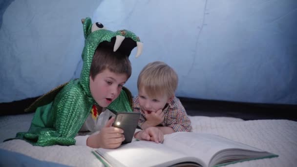 Frère aîné dans un costume de dragon joue avec un plus jeune, les enfants utilisent un téléphone portable avec lampe de poche et lire le livre tout en étant couché dans la tente à la maison — Video
