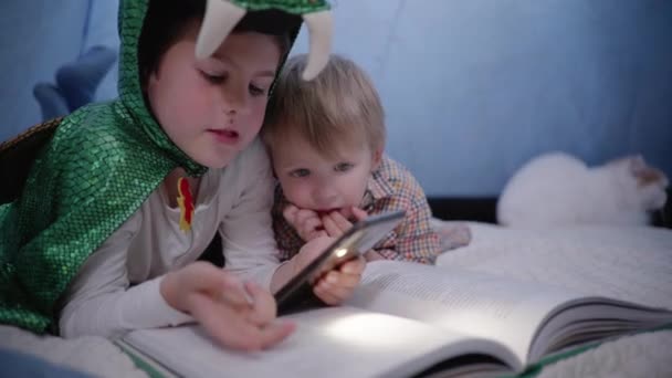 Enfants à la maison, enfant plus âgé en costume de dragon lit livre au jeune frère et utilise gadget avec éclairage dans la tente — Video