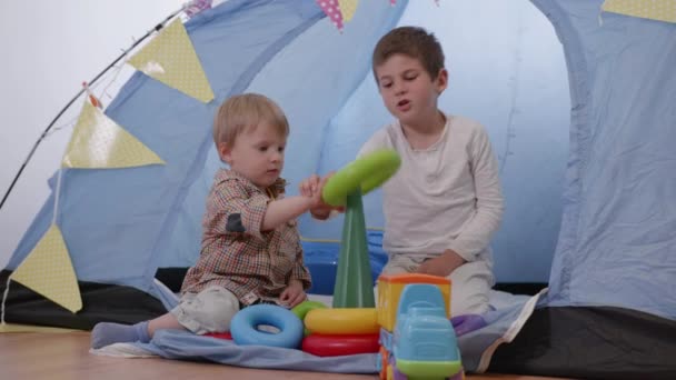 Eigenheimentwicklung, älterer Bruder bringt Jüngeren das Spielen mit Plastikspielzeugpyramide im Zelt bei — Stockvideo