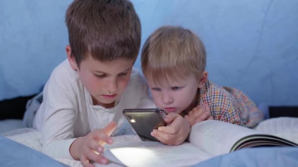 Frère aîné lit des contes de fées pour les plus jeunes et utilise un téléphone avec lampe de poche couché dans une tente à la maison pendant les vacances — Video