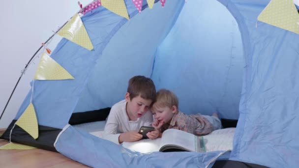 Bästa barndomsvänner, två bröder läser en bok och använder en telefon med ficklampa liggande i ett tält hemma — Stockvideo