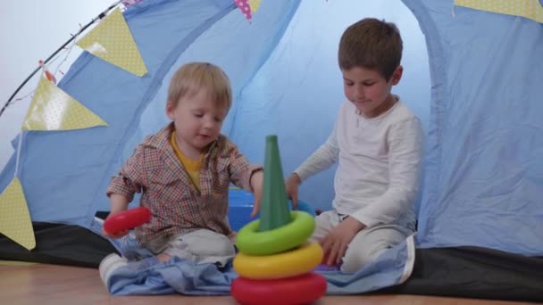 Schlaue kleine Brüder spielen mit einer Spielzeugpyramide aus Plastik, die in Zeitlupe im heimischen Zelt sitzt — Stockvideo