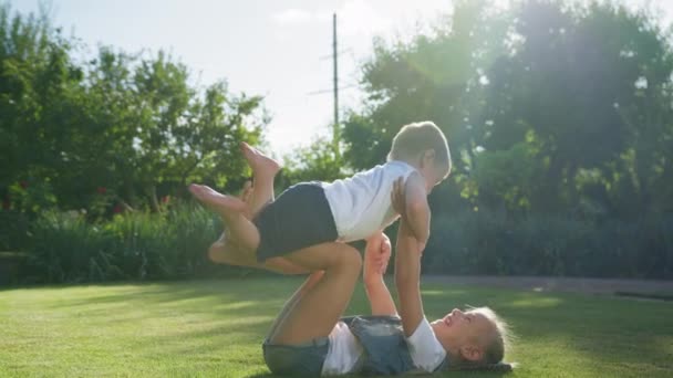 Feliz vacaciones, hermana mayor rollos de hermanito lindo en sus pies acostado en la hierba verde en el parque — Vídeo de stock