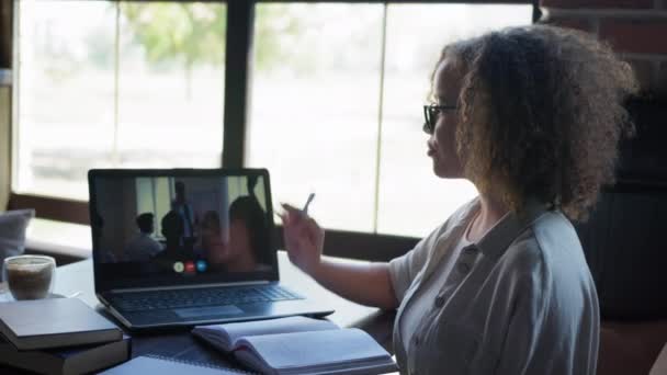 Lecciones en línea, estudiante femenina se entrena de forma remota utilizando tecnologías modernas levanta la mano y toma notas en el cuaderno mientras está sentado con el ordenador portátil en la mesa en el restaurante — Vídeo de stock