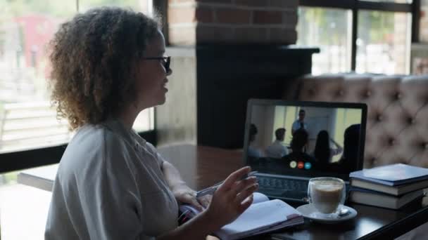 Formation en ligne, femme avec des études de lunettes en ligne lors d'une conférence à l'aide d'un appel vidéo lève la main et prend des notes dans un carnet — Video