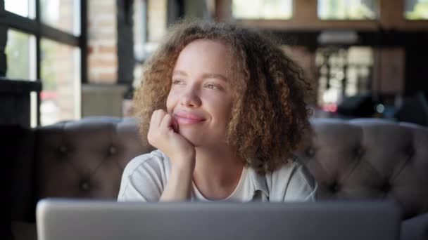 Дівчина дивиться у вікно посміхається і дивиться в камеру, сидячи з ноутбуком в кафе — стокове відео