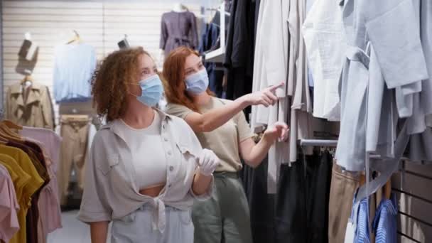 Młode kobiety w maskach medycznych wybierające ubrania na półkach sklepowych — Wideo stockowe