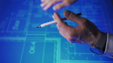 In an office, leaning over a large monitor instead of a desk, a man actively uses an interactive diagram on the screen. The camera focuses on his hand, wearing a watch and holding a pencil. The colors